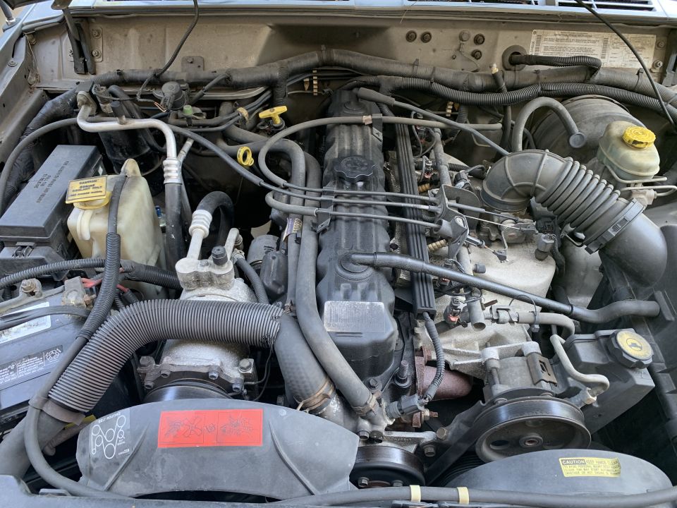2000 Jeep Cherokee engine bay with visible hoses, belts, and engine components. Text on a red label is partially visible