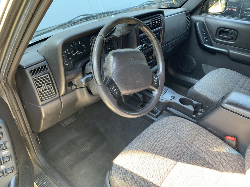 Jeep Cherokee interior with gray patterned seats, dark gray dashboard, and a Jeep steering wheel. The driver's side door panel is visible