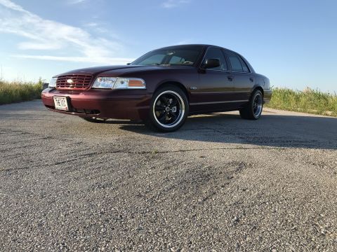 2004 Ford Crown Victoria - Maroon Ford Crown Victoria with aftermarket wheels sits on a gravel road under a blue sky.