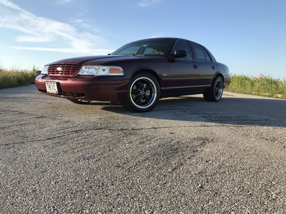 Maroon Ford Crown Victoria with black aftermarket wheels parked on a gravel road