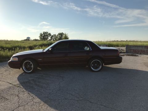 2004 Ford Crown Victoria - Side view of a maroon Ford Crown Victoria with aftermarket black rims under a clear sky.