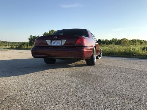 2004 Ford Crown Victoria - Rear view of a maroon sedan with custom wheels, parked outdoors under a clear blue sky.