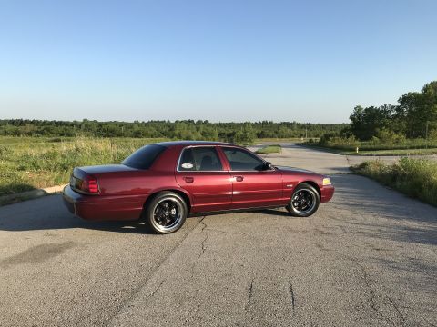 2004 Ford Crown Victoria - A red 2004 Ford Crown Victoria sedan is seen from a three-quarter front angle on a paved road with a wooded background.