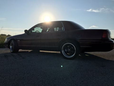 2004 Ford Crown Victoria - A dark 2004 Ford Crown Victoria is captured from the side, custom black wheels under a bright sky.