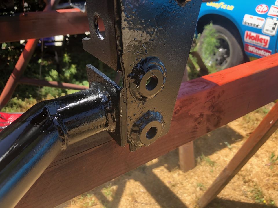 Black painted metal bracket with bolts, attached to red wooden beams. A blue race car is visible in the background