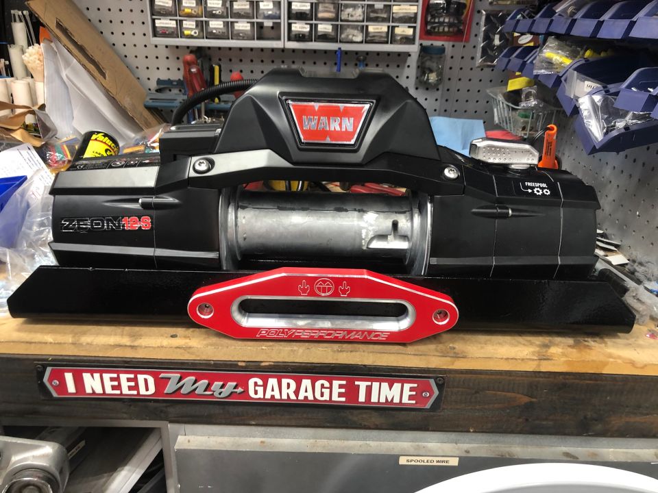 Warn winch mounted on a black base, with a red "Poly Performance" shackle mount in front. "I NEED My GARAGE TIME" sign below
