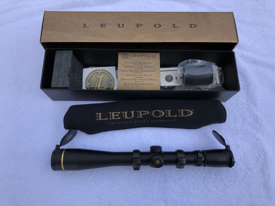Black Leupold rifle scope with gold accents, in its box with accessories and a protective cover