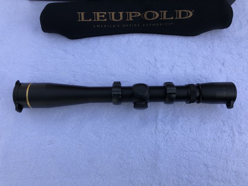 Black Leupold rifle scope with gold accents, mounted on rings, with a black lens cover behind it