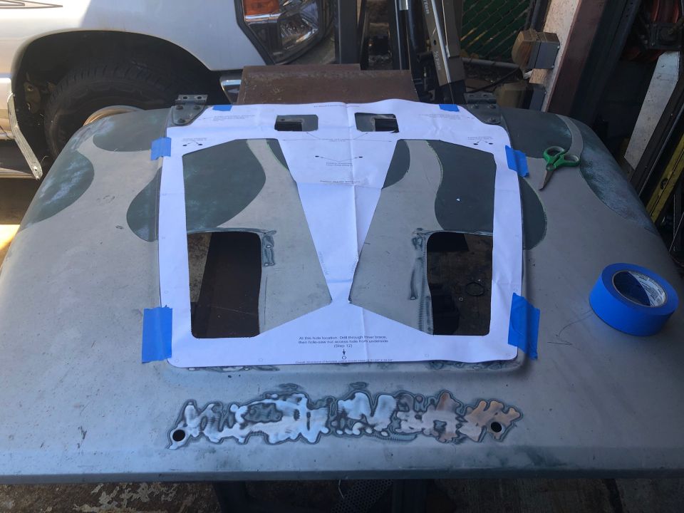 Hood of a vehicle with a paper template taped on it, showing cutouts. Blue tape and scissors are nearby