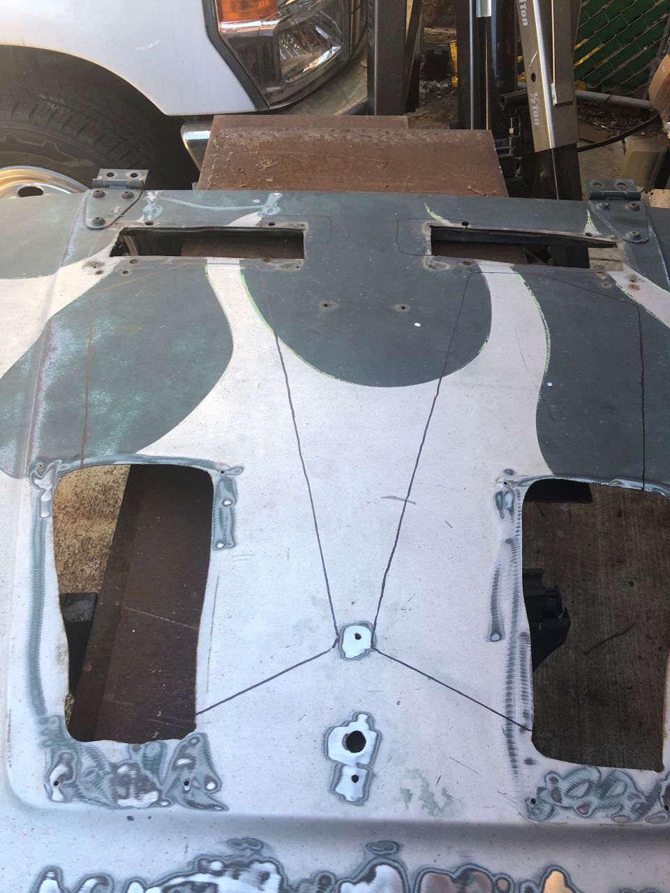 Hood of a Jeep with cutouts and markings for modification. A white truck is partially visible in the background