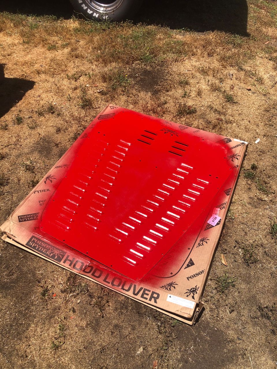 Red hood louver part on a cardboard box. Text on box says 'Poison Spyder Hood Louver'. Part of a tire visible at top left