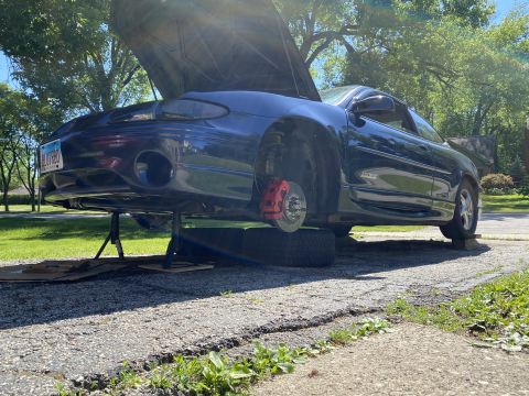 Dark blue Pontiac Grand Prix with hood open, red brake calipers visible, jacked up in a driveway.