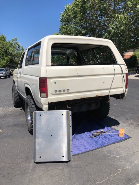 1981 Ford Bronco - The rear of a white 1980s Ford Bronco is shown parked on an asphalt surface. A large, flat metal skid plate sits on the ground in front of…