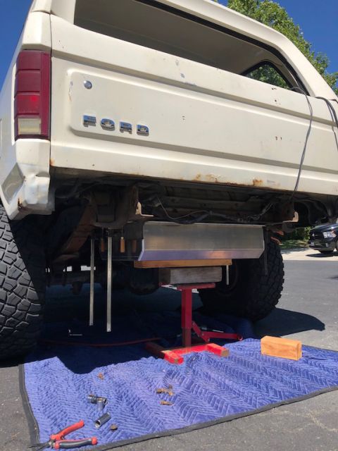 1981 Ford Bronco - The rear underside of a white 1980s Ford Bronco is shown with a new aluminum fuel tank being installed. Tools and a floor jack are laid out…