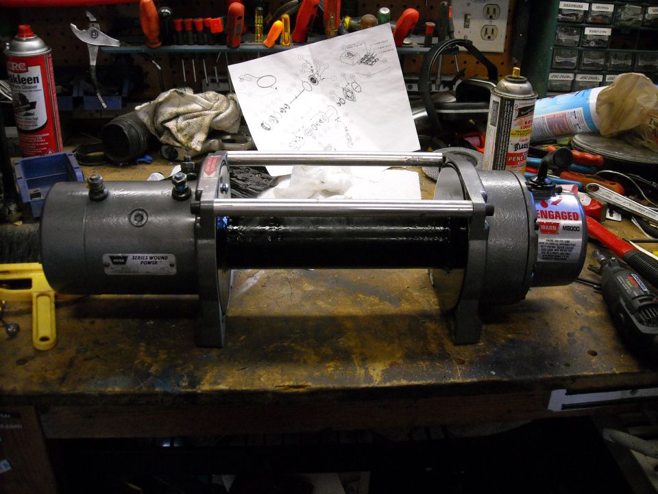 Gray electric winch with black drum, labeled WARN MB8000, on a workbench with tools.
