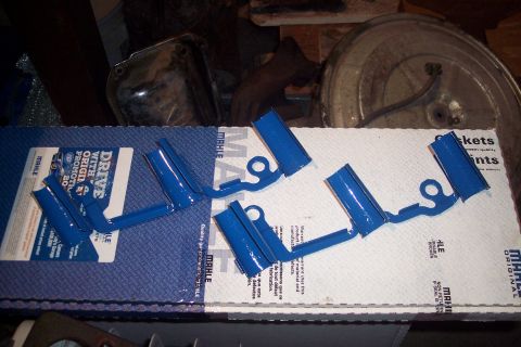 1978 Ford Bronco - Four blue metal brackets with circular holes are laid out on top of a white cardboard box with Mahle branding. A promotional card with the…