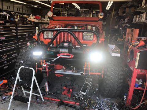 1992 Jeep Wrangler - An orange Jeep Wrangler is parked in a garage with its front end elevated on jack stands, surrounded by tools and equipment. The vehicle fe…