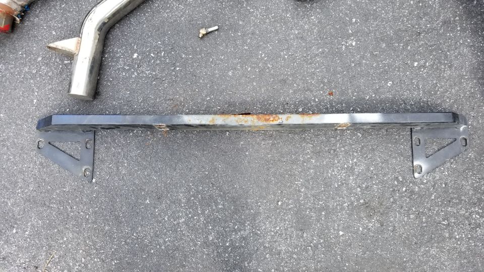Rusty bumper bar with mounting brackets on asphalt, partial chrome exhaust pipe.