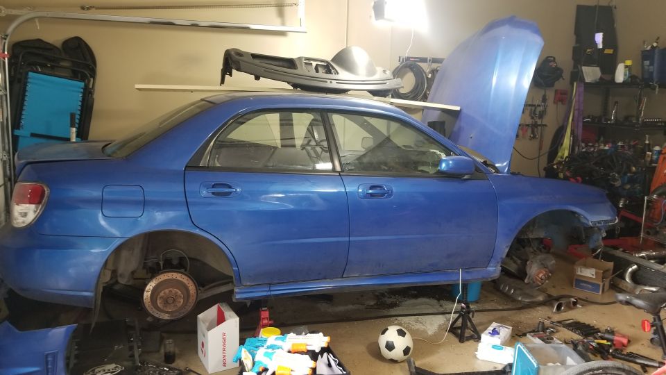 Blue Subaru Impreza sedan in a garage, hood up, front end disassembled, on jack stands.