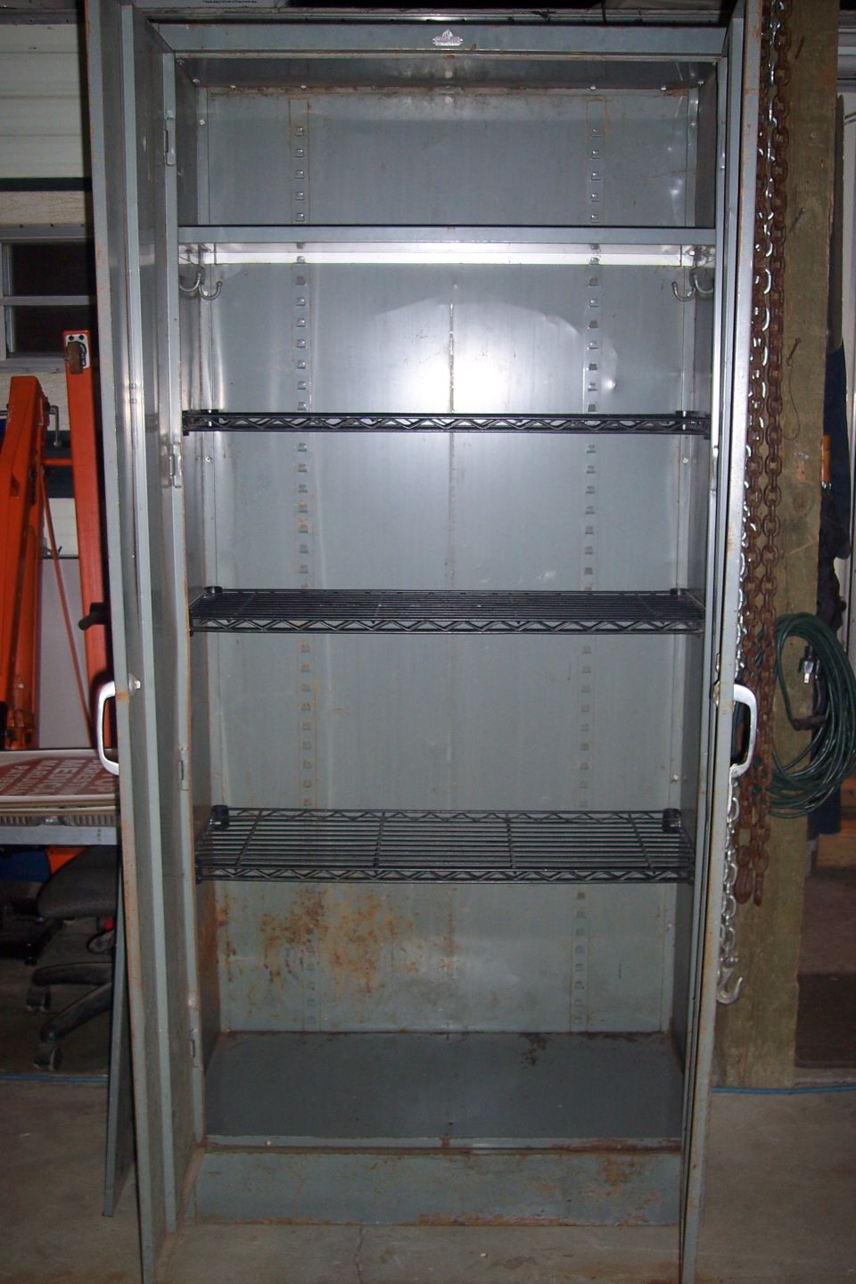 Empty gray metal cabinet with three wire shelves, showing rust and wear.