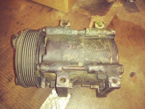 1996 Ford Bronco - A dirty, black AC compressor with a pulley is shown resting on a wooden surface. The compressor has mounting brackets and appears to be a v…