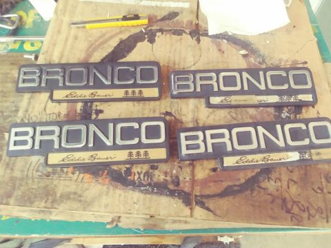 1996 Ford Bronco - Four Ford Bronco Eddie Bauer emblems are laid out on a wooden surface. The emblems feature silver lettering for "BRONCO" and gold backing w…