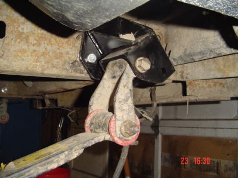1989 Ford Bronco - Underside view of a black bracket/mount welded to the frame, with a rusty leaf spring or radius arm attached via a large bolt through a red…