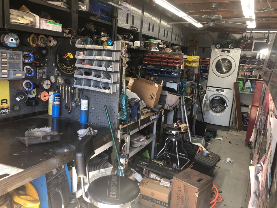 Cluttered garage scene with workbench, tools, storage bins, and appliances.