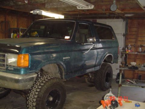 1989 Ford Bronco - A green two-door Ford Bronco SUV with a suspension lift and large off-road mud tires on black steel wheels, parked in a wood-paneled garage…