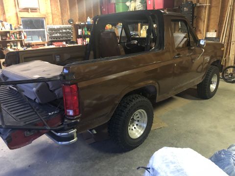 1980 Ford Bronco - The rear passenger side of a brown 1980 Ford Bronco is shown in a garage. The vehicle has a removable top, aftermarket wheels, and a spare…