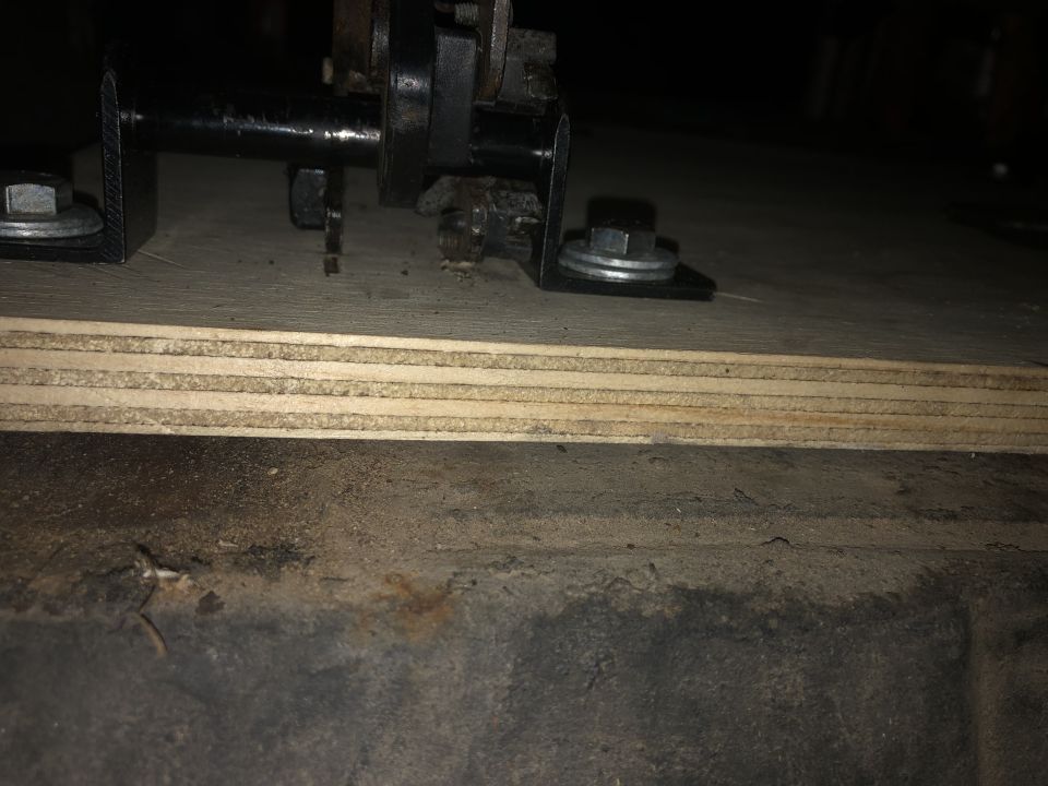 Metal bracket with bolt and washer on plywood, above vehicle undercarriage.