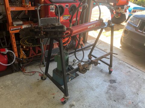 1992 Jeep Wrangler - A red hydraulic tube bender is mounted on a black metal stand with wheels. The bender is in a garage setting with shelves and other equipme…