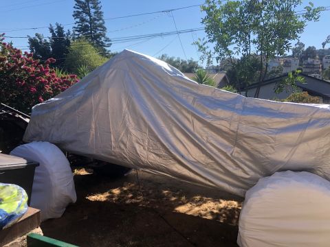 1992 Jeep Wrangler - A tarp covers a vehicle, with two large white-wrapped tires visible on the left and right sides. The vehicle is parked outdoors in a yard w…