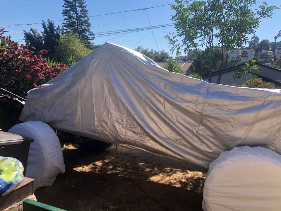 Vehicle covered by a tarp with two large white-wrapped tires visible.