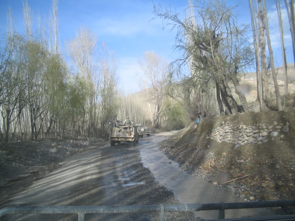 Military vehicles driving down a dirt road lined with trees. Some people are visible on the side of the road