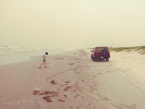 1996 Ford Bronco - A red Ford Bronco is parked on a sandy beach next to the ocean, with a young boy walking in the foreground.