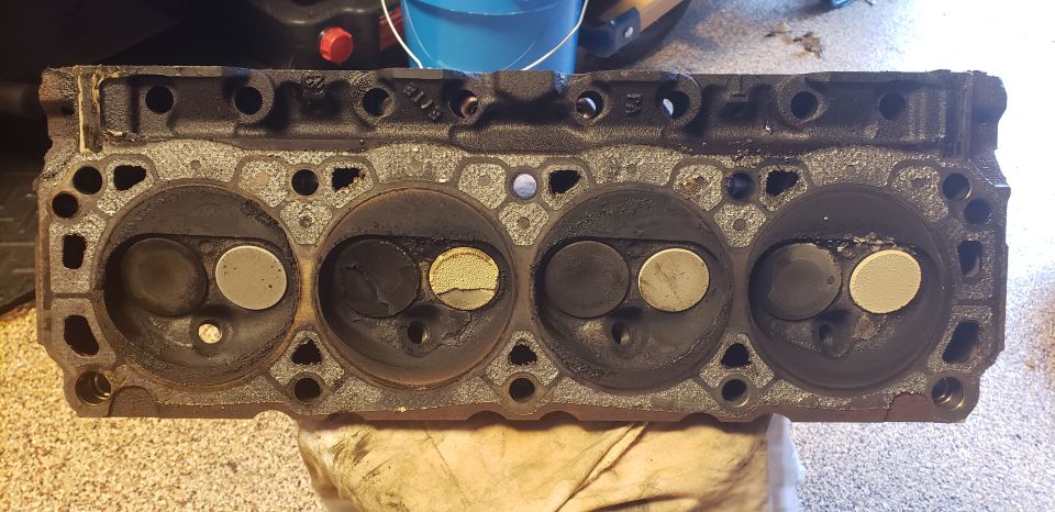 Deck side of a dirty cast-iron cylinder head showing four carbon-covered combustion chambers and valves