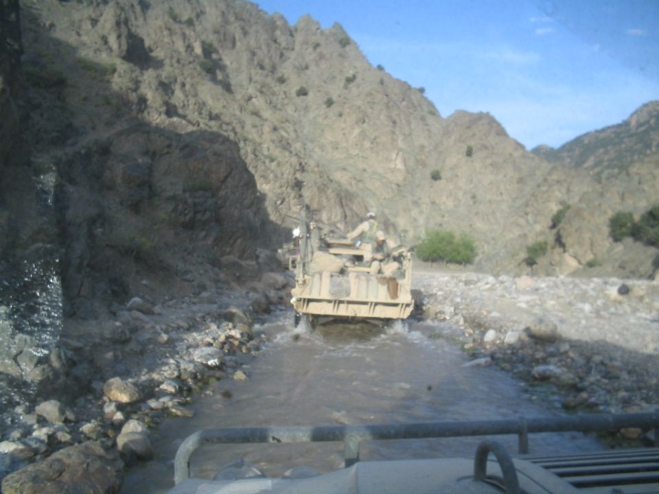Tan military vehicle with soldiers driving through a shallow stream in a rocky, mountainous area