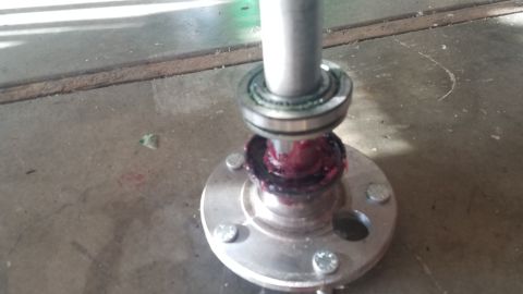 1985 Ford Bronco - A close-up view shows a metal shaft with a bearing being greased with red lithium grease, positioned above a silver wheel hub with four bol…