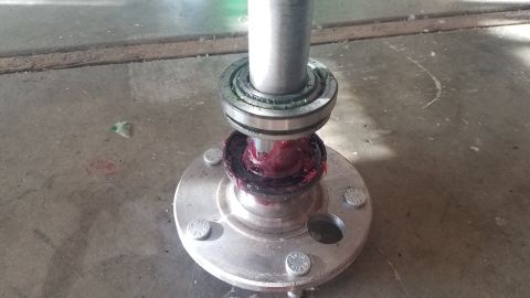 1985 Ford Bronco - A metal shaft is being greased with red lithium grease as it sits on a silver wheel hub with four bolts. The scene appears to be on a concr…