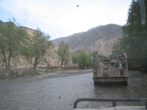1981 Ford Bronco - A military vehicle carrying soldiers navigates through a shallow river with mountainous terrain and buildings visible in the distance.