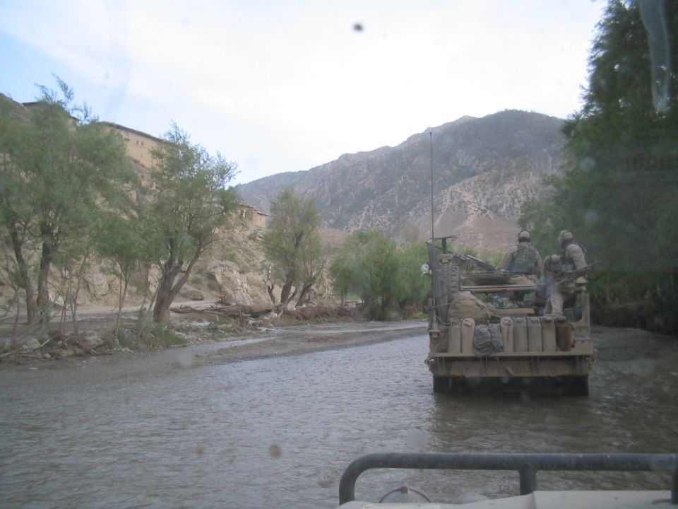 Military vehicle with soldiers driving through a river, mountains in background