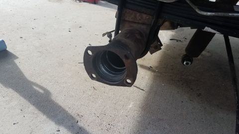 1985 Ford Bronco - A close-up view of a rusty exhaust pipe flange attached to a vehicle's leaf spring suspension system. The flange has four bolt holes and sh…
