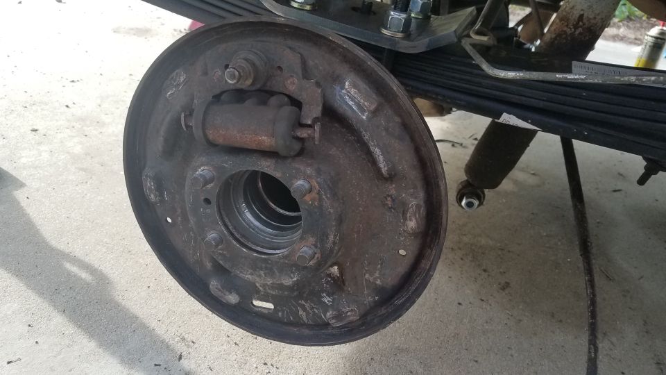 Rusty brake drum assembly with wheel cylinder and brake shoes. Leaf springs and shock absorber in background.