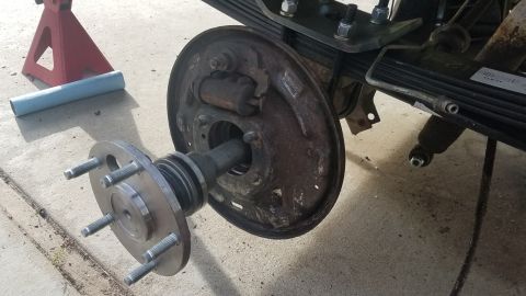 1985 Ford Bronco - A close-up view of a vehicle's rear axle assembly, showing the brake drum, hub with wheel studs, and part of the leaf spring suspension sys…
