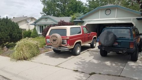 1985 Ford Bronco - A red 1985 Ford Bronco with a tan top is parked on a driveway next to a teal Jeep Cherokee. Both vehicles are parked in front of a suburban…