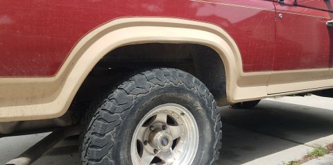 1985 Ford Bronco - Close-up side view of the rear fender and wheel well of a red and tan Ford Bronco. The vehicle has an aftermarket off-road tire and a chrom…