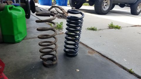 1985 Ford Bronco - Two coil springs are shown on a concrete surface. The spring on the left is older and rusty, while the spring on the right is new and black…
