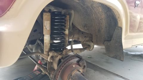 1985 Ford Bronco - The driver's side front suspension of a vehicle is shown, featuring a new black coil spring and a worn shock absorber. A red hydraulic jack…