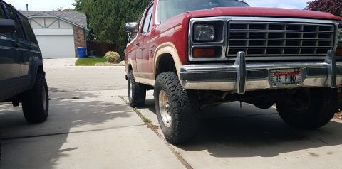 1985 Ford Bronco - The front quarter view of a red and tan 1980s Ford Bronco is parked on a concrete driveway next to a dark colored SUV. The Bronco has a chr…